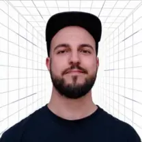 Portrait of a man with a dark beard wearing a black cap, standing in a white grid-lined tunnel backdrop.