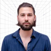 Portrait of a man with a beard and shoulder-length hair, wearing a blue denim shirt, facing the camera against a white grid background.